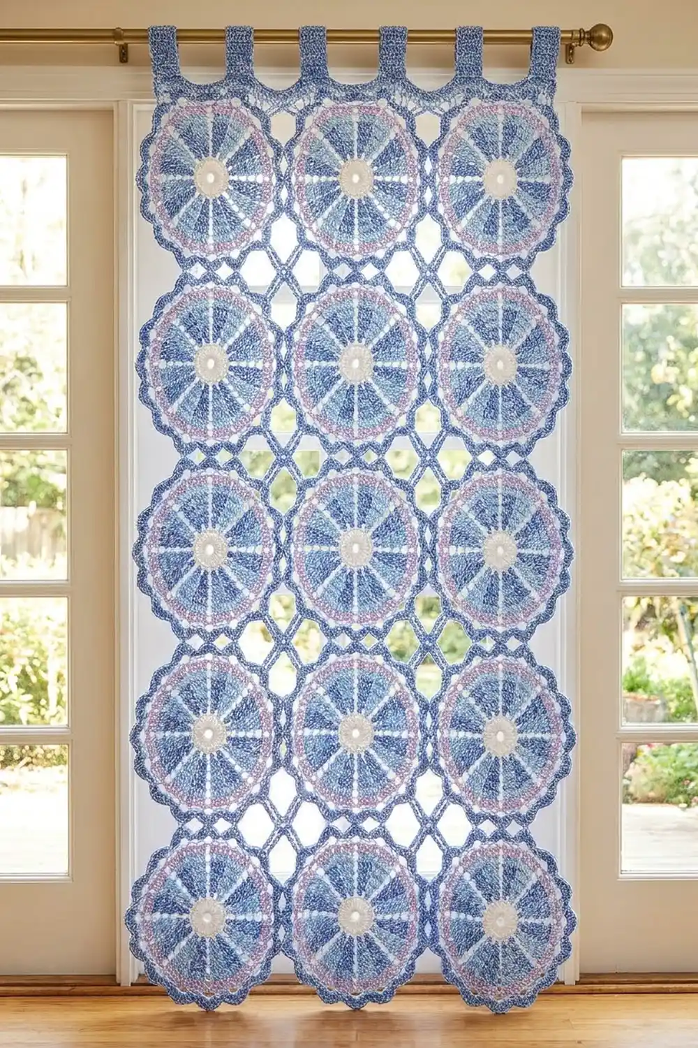 Blue and dusty pink crocheted curtain with cream medallion centers hangs on a brass rod over a sunny garden window.