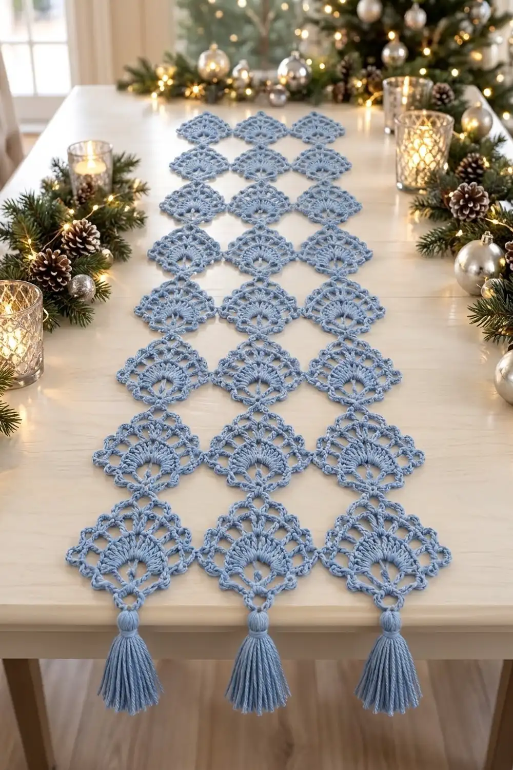 Light blue crocheted table runner with diamond lace and tassels displayed on white table with holiday garland decorations.