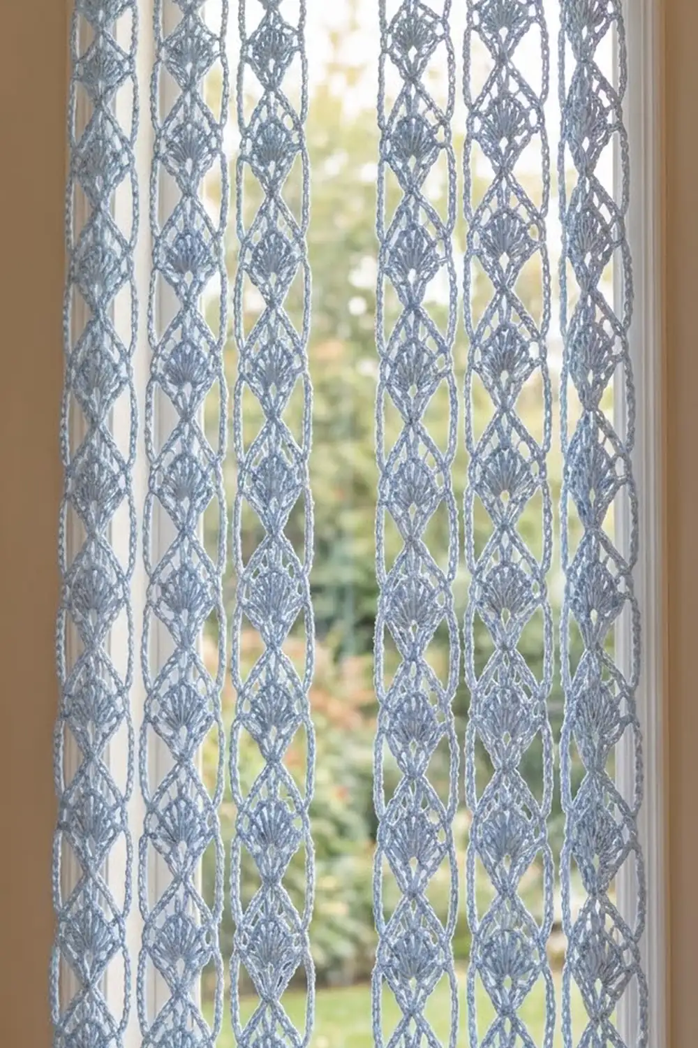 Gray and cream crocheted lace curtains with diamond patterns hang at a window overlooking green outdoor views.