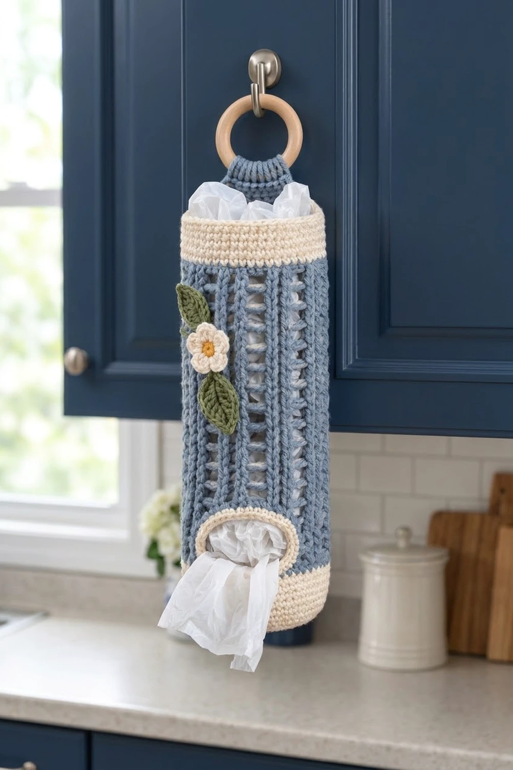 Slate-blue crocheted tissue holder with cream bands and white flower detail displayed on a modern kitchen counter.