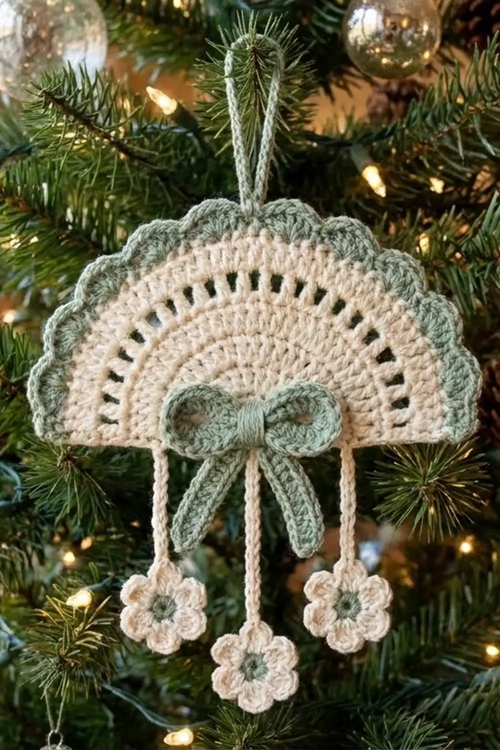 Cream and sage green crocheted fan ornament with bow and flowers, hanging on a glowing Christmas tree with evergreen branches.