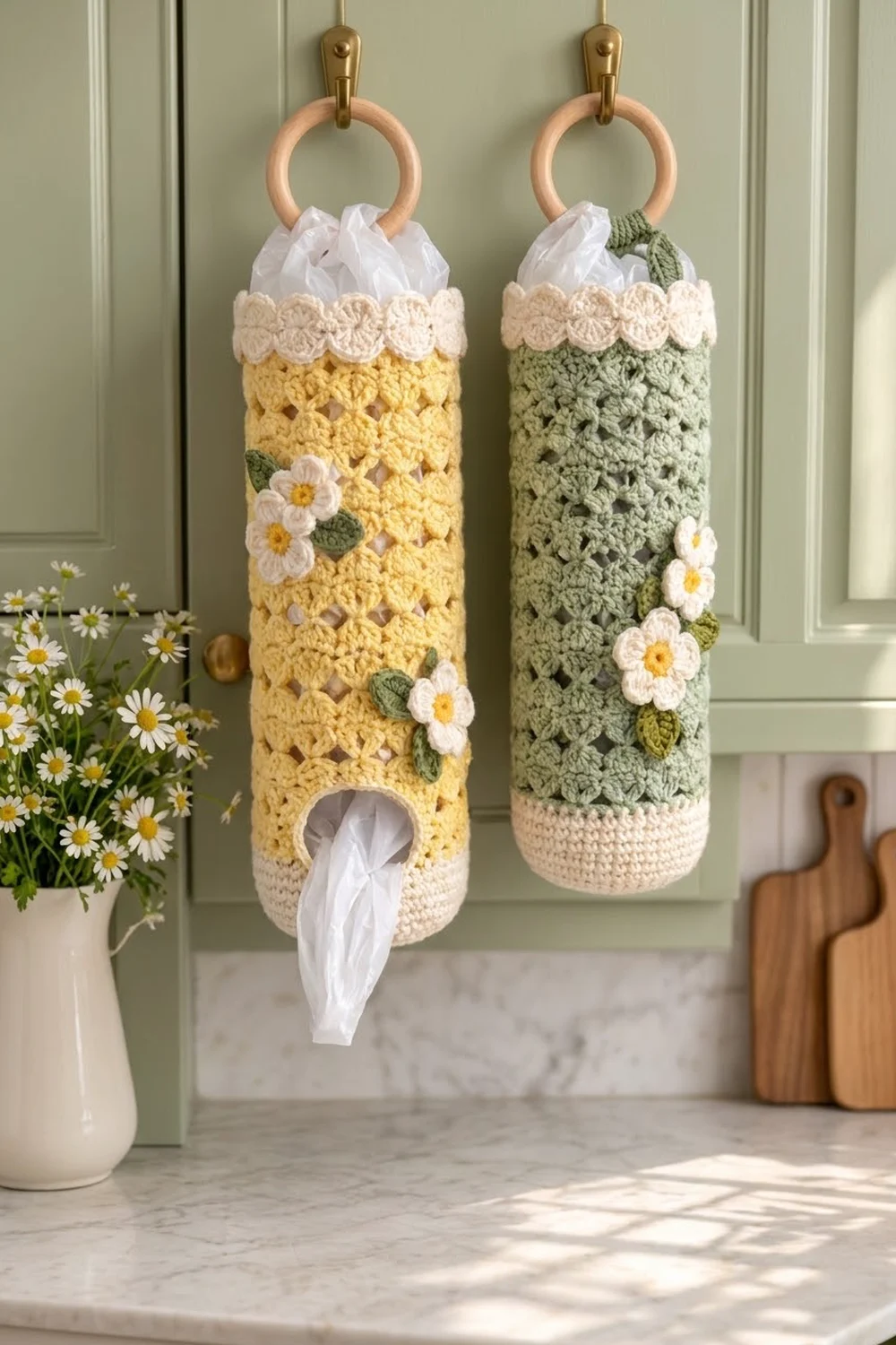 Crocheted tissue holders in yellow and sage green adorned with white flowers hang on a kitchen cabinet.