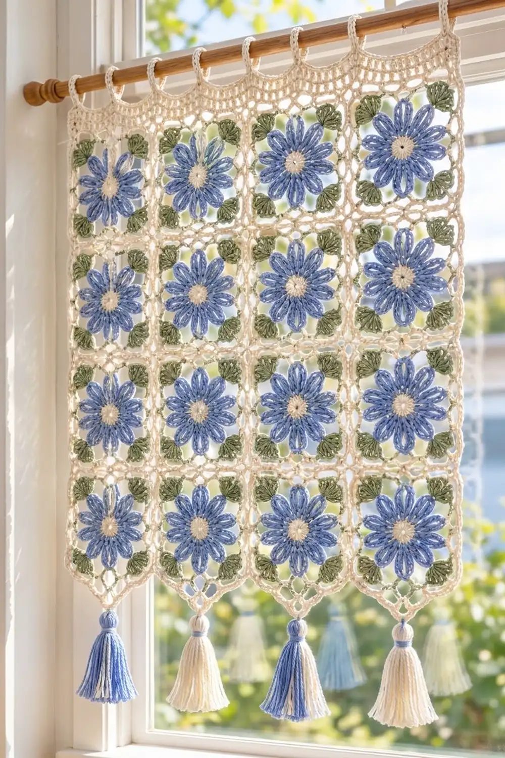 Cream-colored crocheted floral valance with blue daisies and green leaves hanging from a wooden rod at a sunlit window.