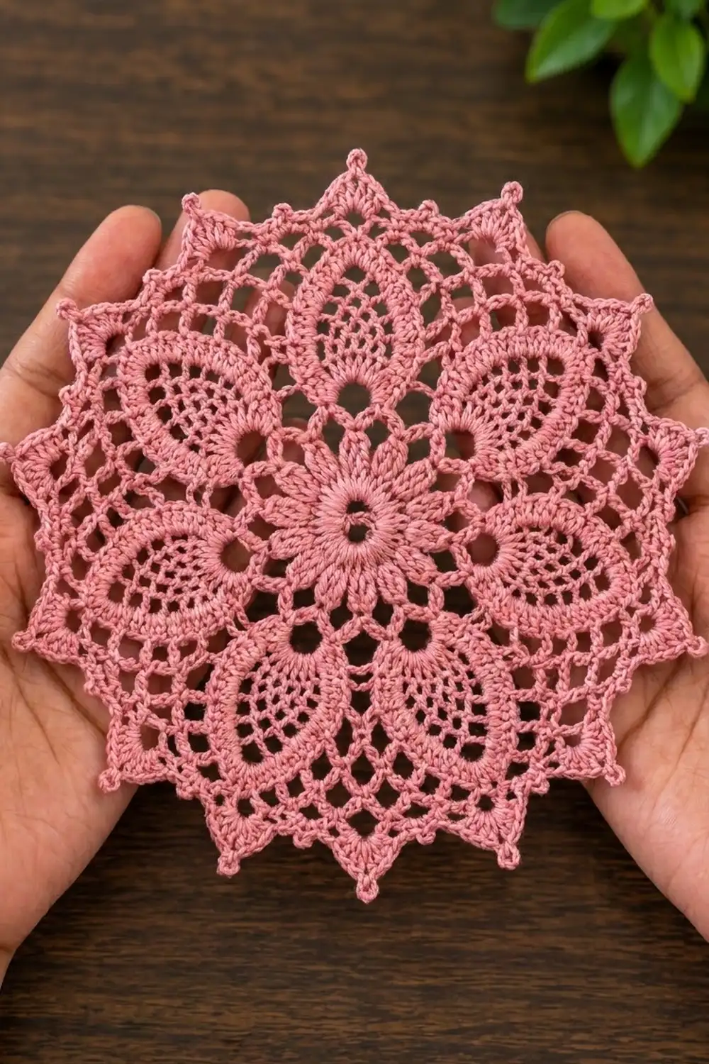 Dusty rose crocheted doily with eight-pointed mandala design held in hands above a dark wooden table with green foliage visible.