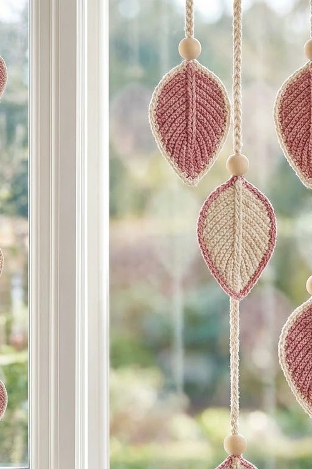 Crochet leaf ornaments in cream and dusty rose hanging from braided cords with wooden beads in a sunlit window.