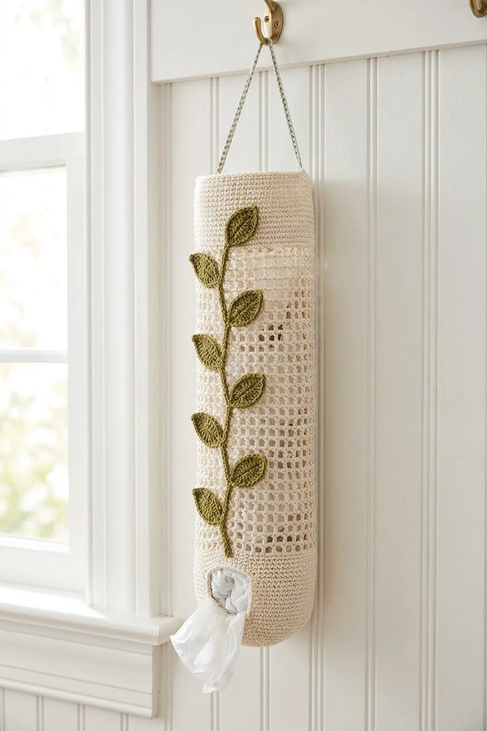 Cream-colored crocheted tissue holder with olive leaves hanging from brass hook against white paneled wall.