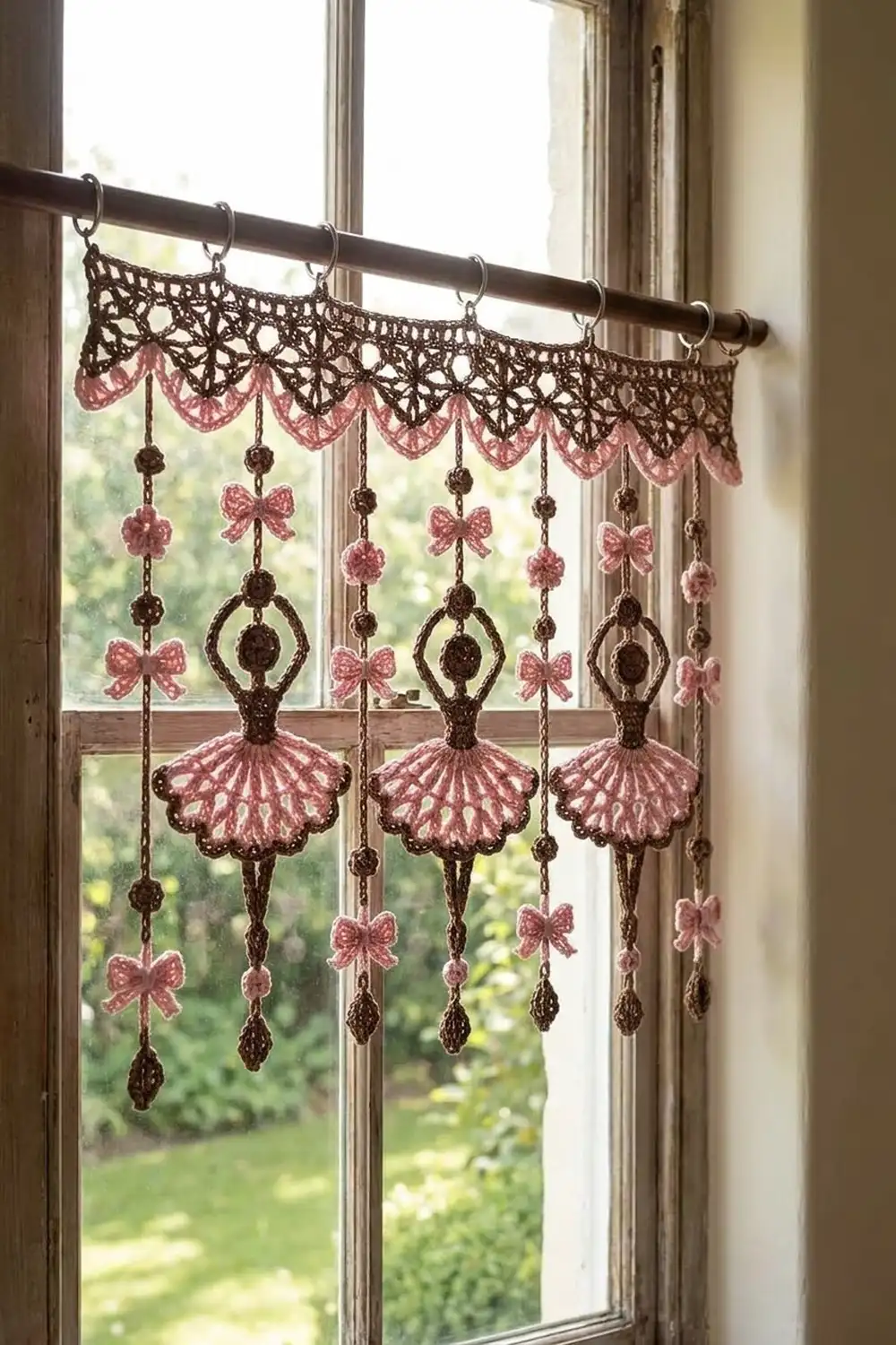Crocheted pink and brown ballet-themed window decoration hanging in front of a bright window with green garden view.