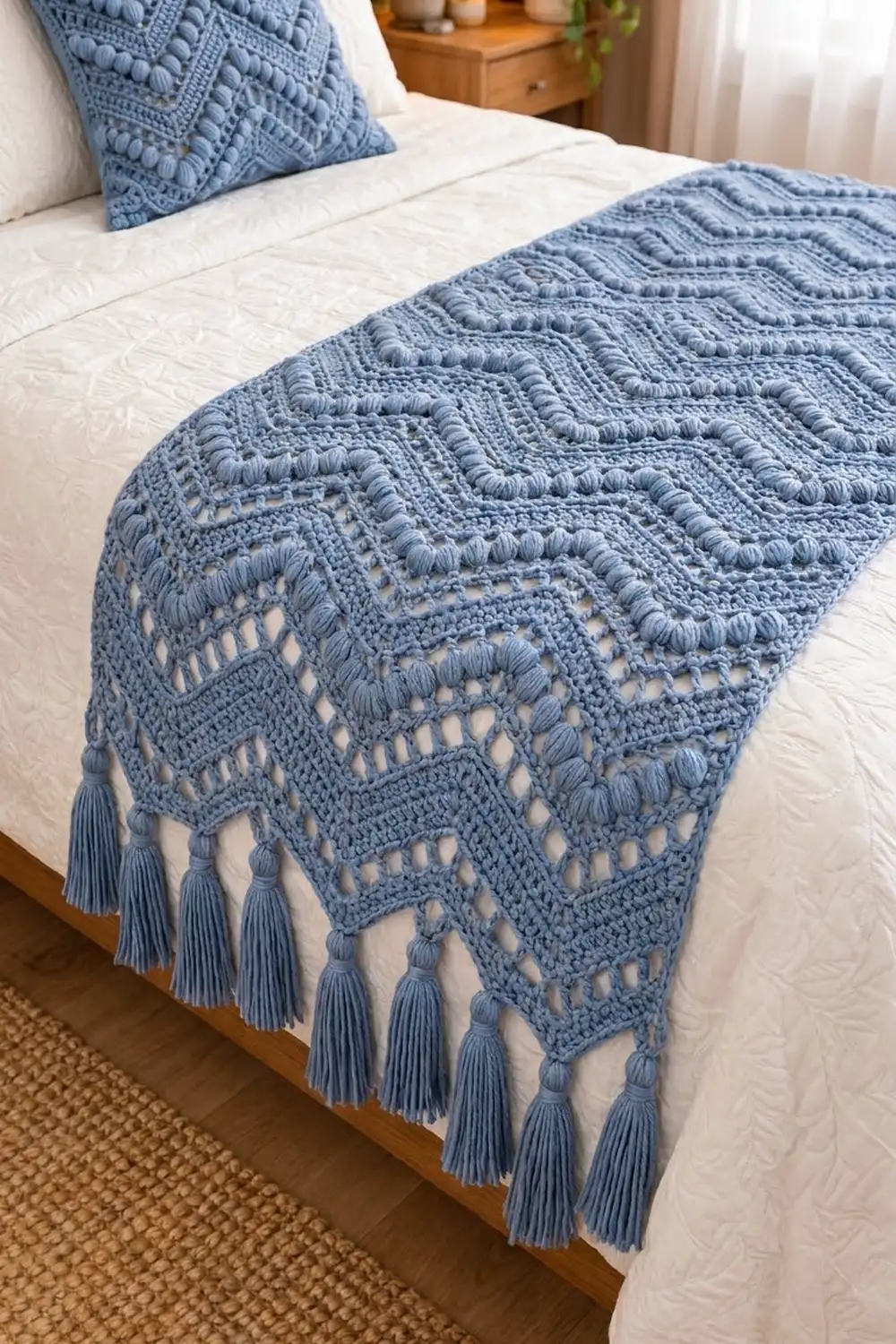 Dusty blue crocheted blanket with intricate lace patterns and fringe trim arranged on white bedding in natural sunlight.