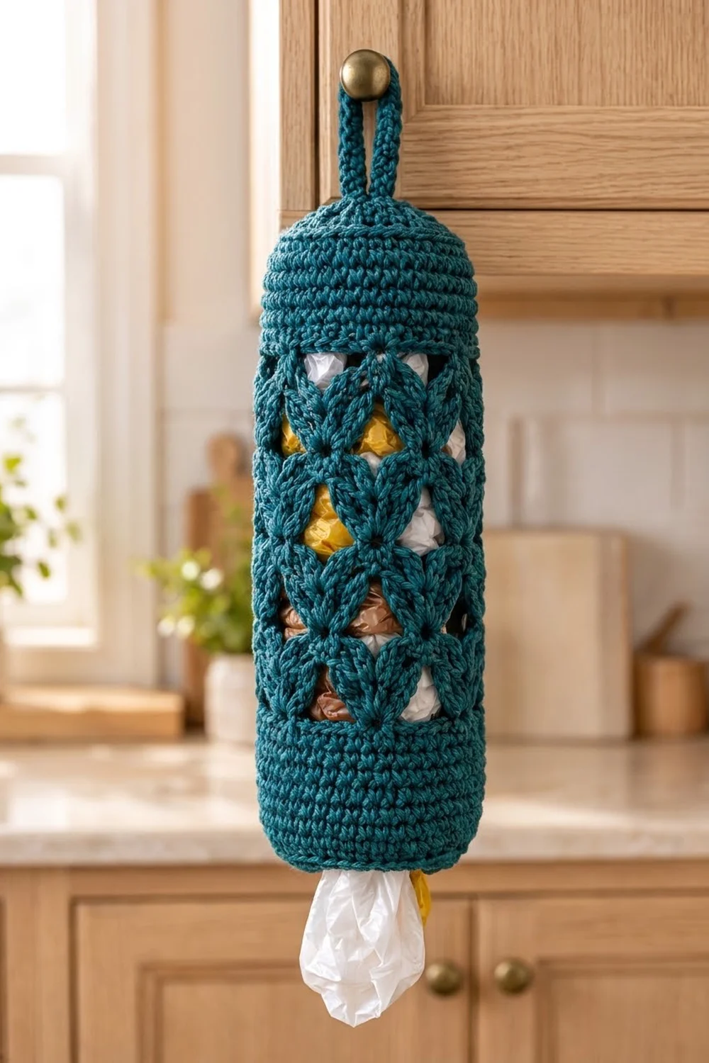 Teal crocheted plastic bag holder with brass knob and lattice weave displayed on wooden kitchen cabinet.