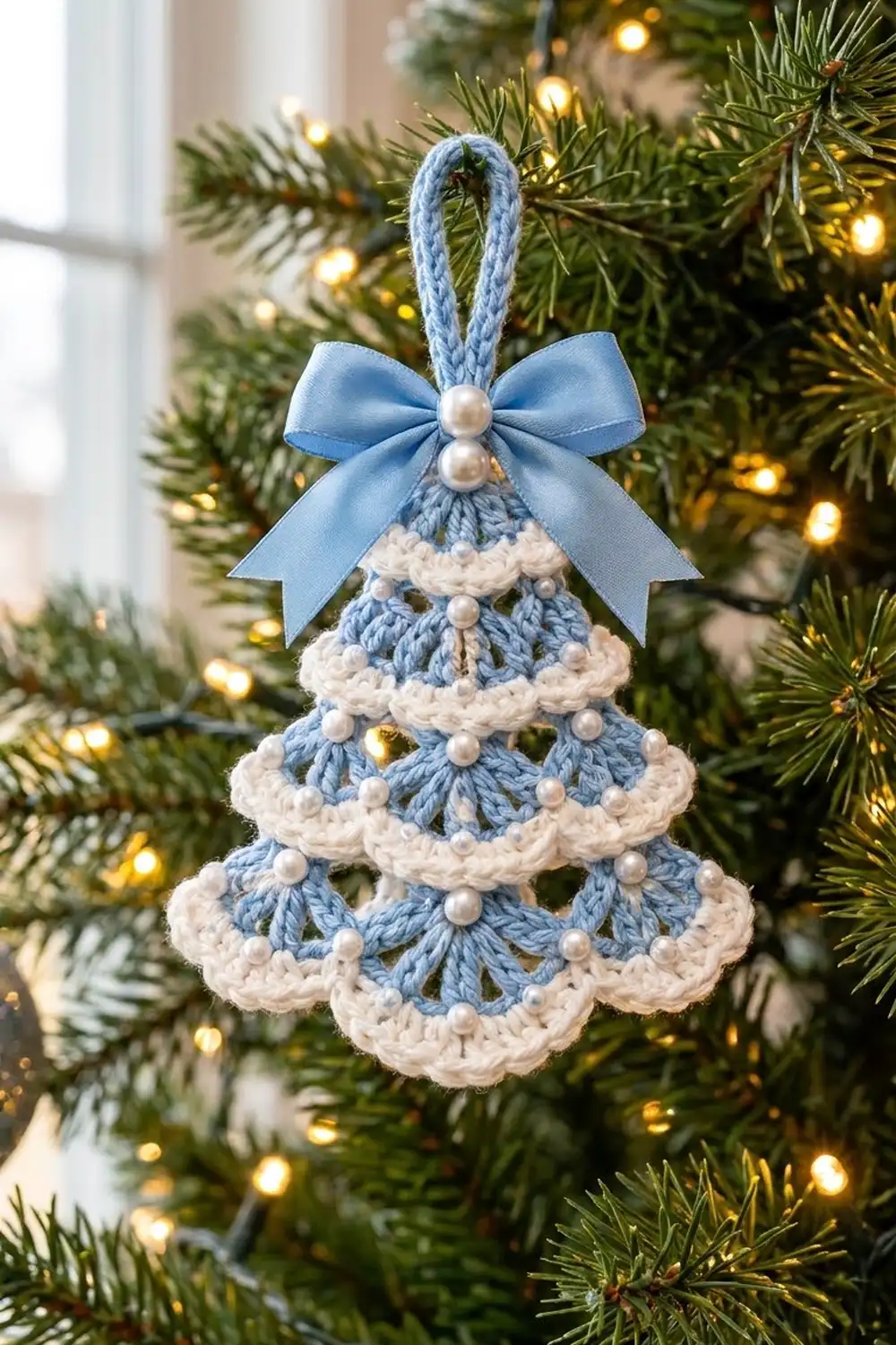 Blue and cream crocheted Christmas tree ornament with pearl beads and bow, hanging on a lit evergreen tree.
