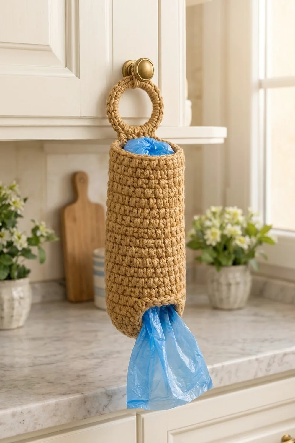 Tan crocheted bag holder with rope handle and brass knob stores blue plastic bags on a sunny kitchen counter.