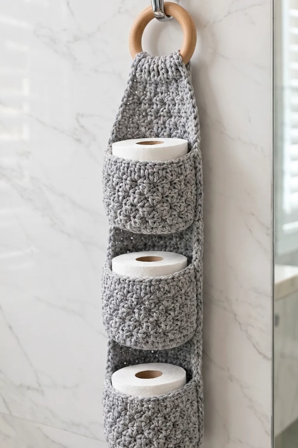 Gray crocheted toilet paper holder with wooden ring hanging on a marble wall, holding three white paper rolls.