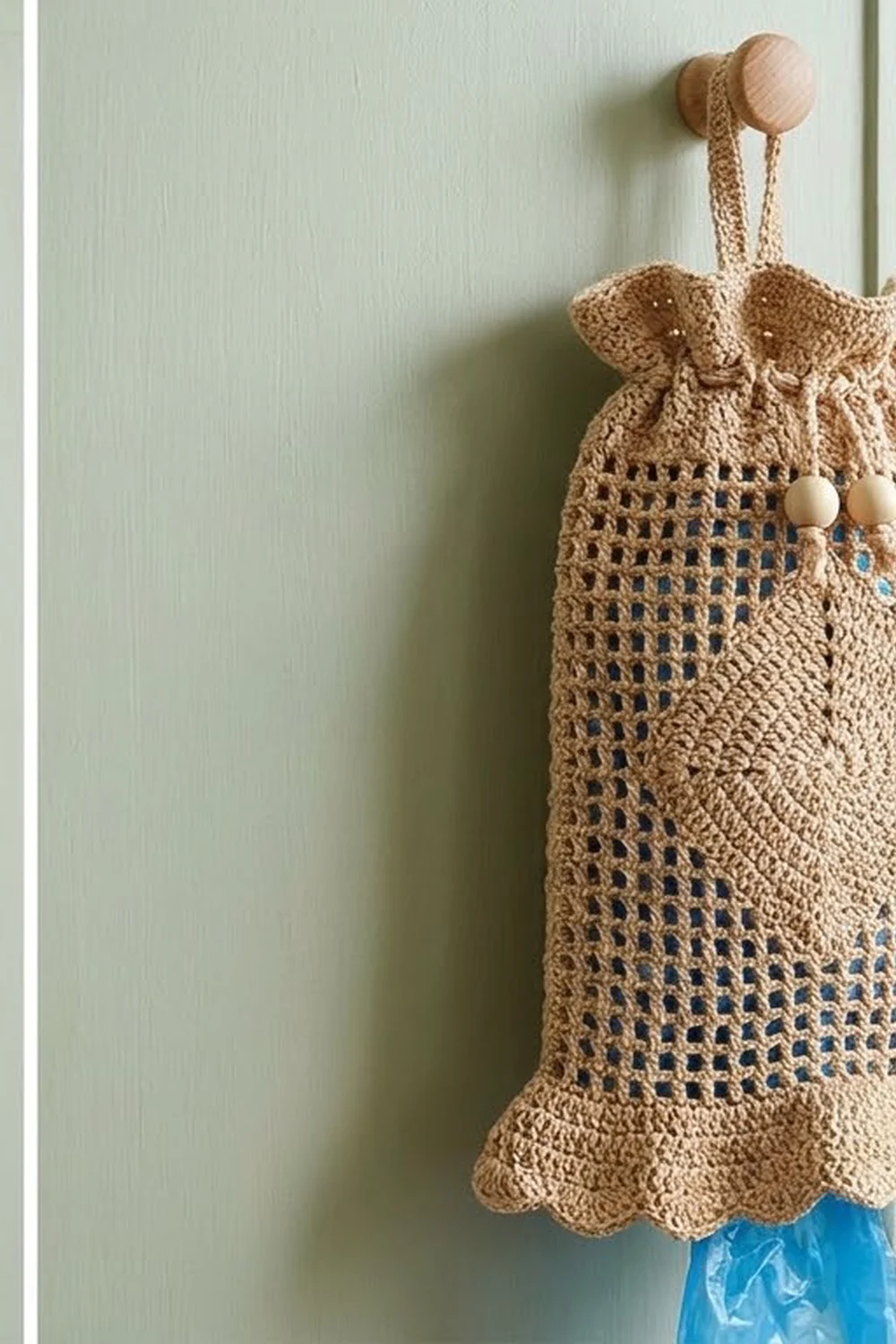 Crocheted tan storage pouch with wooden handles and beaded details hanging on pale green wall.