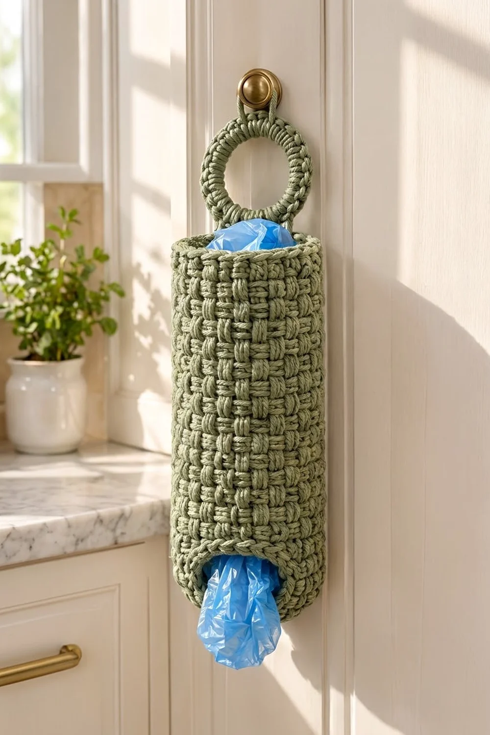 Sage green crocheted bag holder with brass ring mounted on white cabinet beside a potted plant in sunlit kitchen.