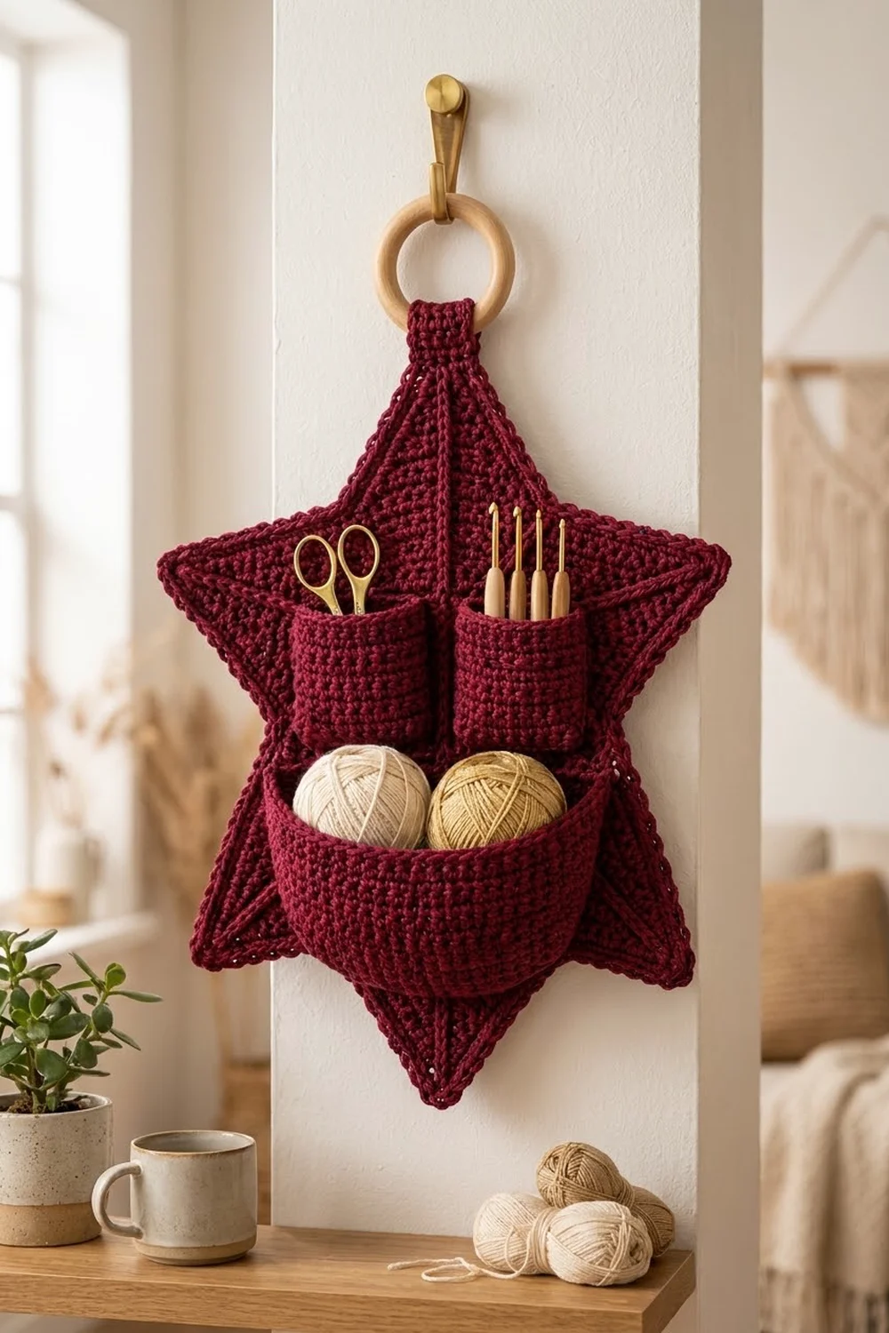 Deep burgundy crocheted star organizer with wooden ring hanging on a white wall, holding yarn and crochet tools in its pockets.