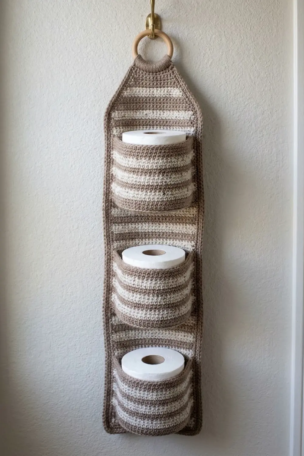 Striped tan and cream crocheted toilet paper holder hanging on a white wall, holding three white paper rolls.