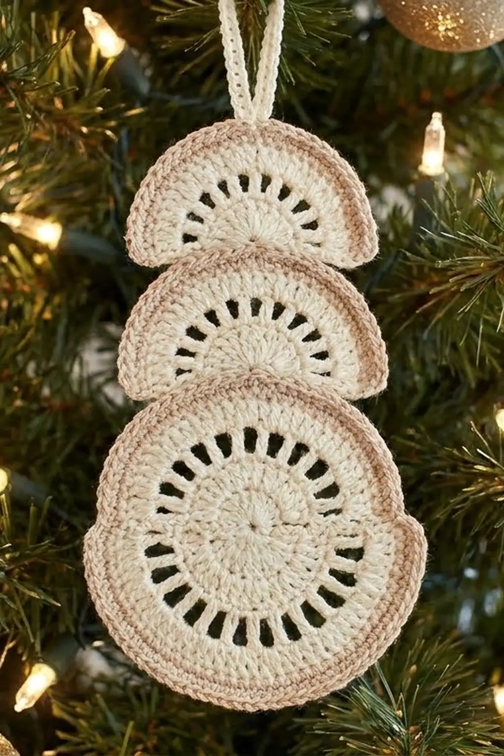 Cream crochet ornament with three stacked rounds and black details hanging on evergreen tree with lights.
