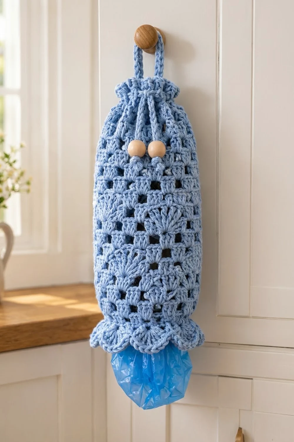 Blue crocheted bag holder with wooden bead eyes, holding a plastic bag, hung on white wall paneling in sunlight.