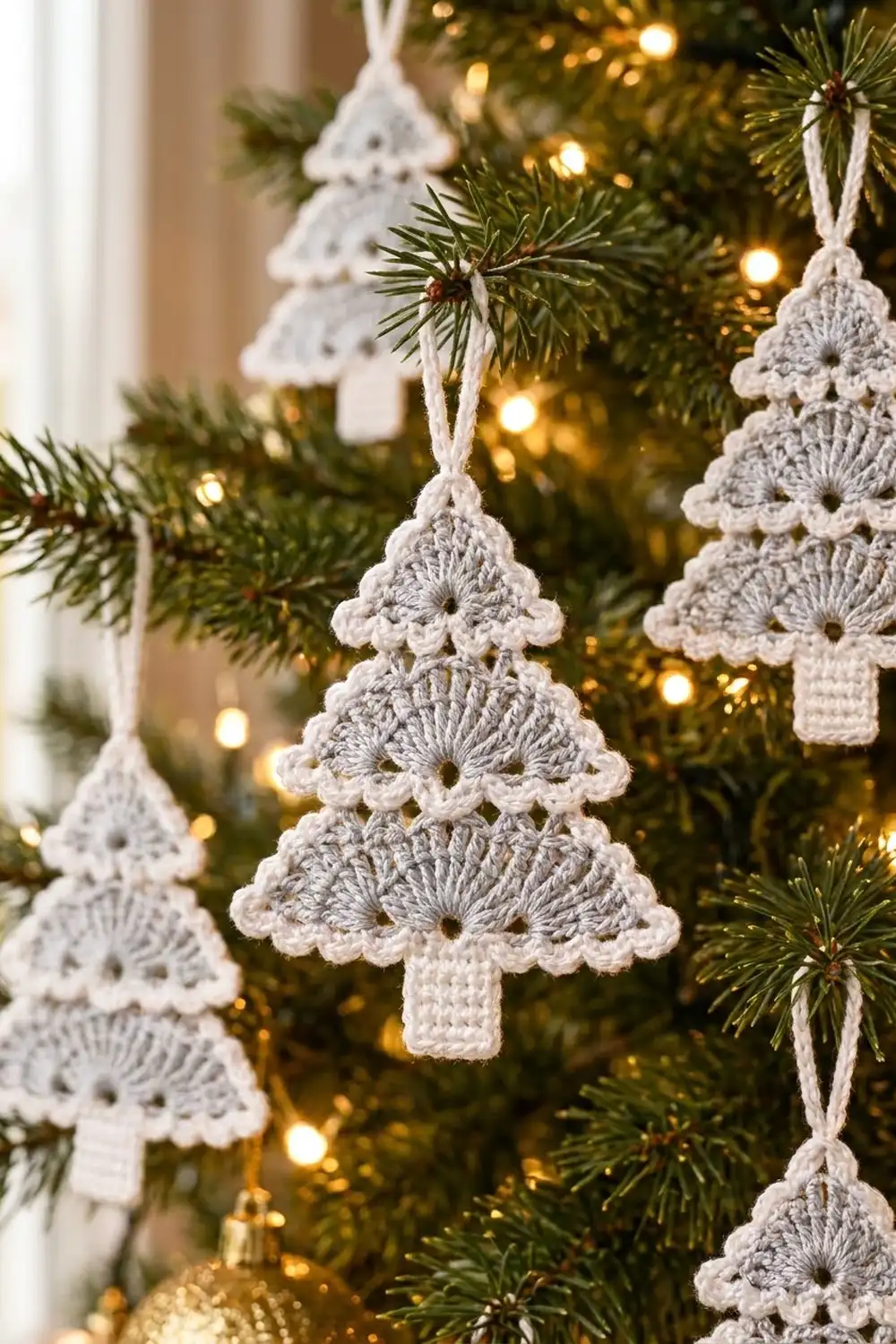 Gray crocheted tree ornaments with white ribbon hangers displayed on an evergreen tree with warm golden lights.