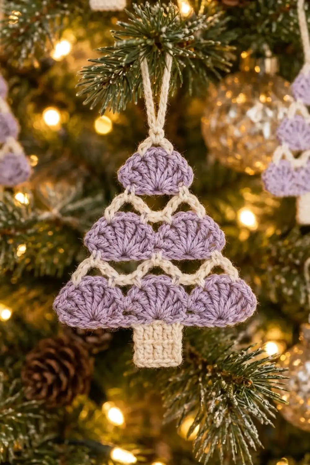 Lavender-and-cream crocheted Christmas tree ornament hanging from ribbon on an evergreen tree with warm golden lights.