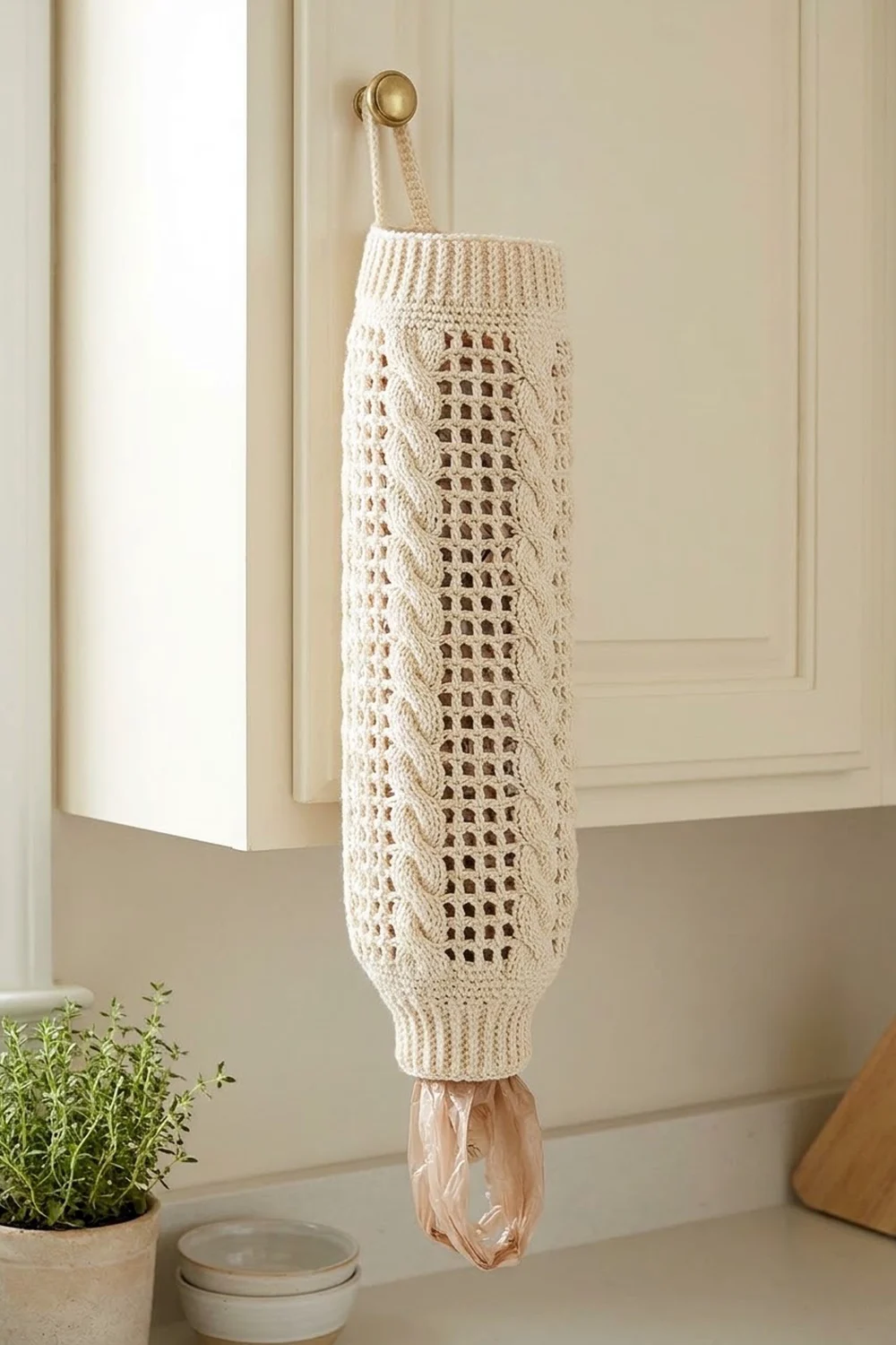White crocheted bag dispenser with lace patterns hanging from brass hook on neutral beige wall.