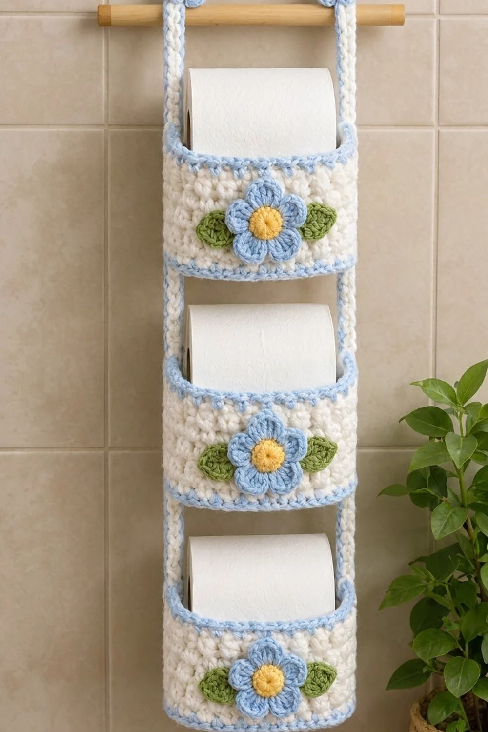 Cream crocheted toilet paper holder with light blue flower appliqués hanging on a bathroom tile wall.