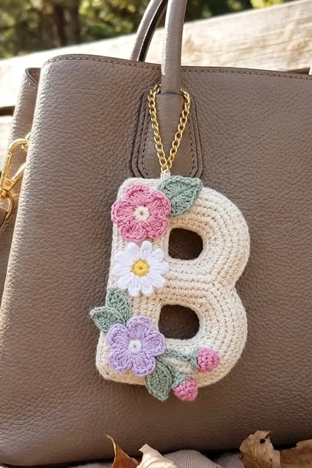 Cream-colored crocheted B-shaped keychain with pink, white, and purple flowers on a gray leather handbag outdoors.