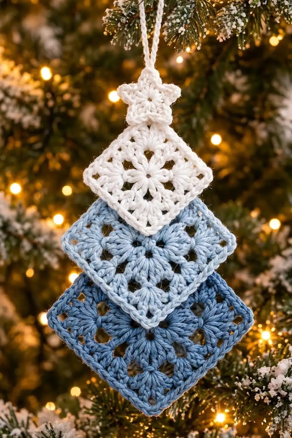 Crocheted tree ornament with white star top, white middle square, and blue bottom squares on snowy evergreen with warm lights.