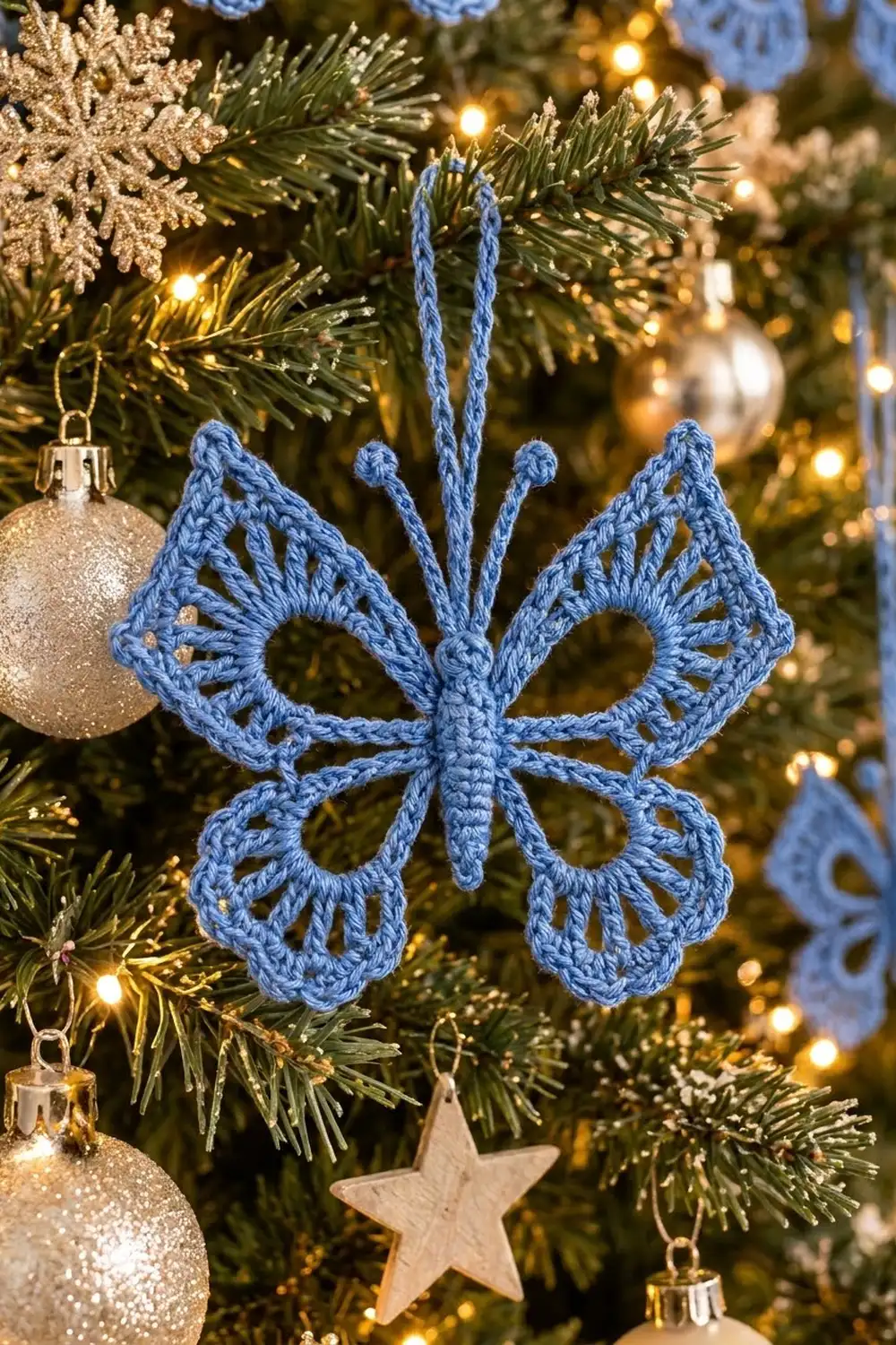 Blue crocheted butterfly ornament hanging on a Christmas tree with warm lights and gold glitter ball ornaments.