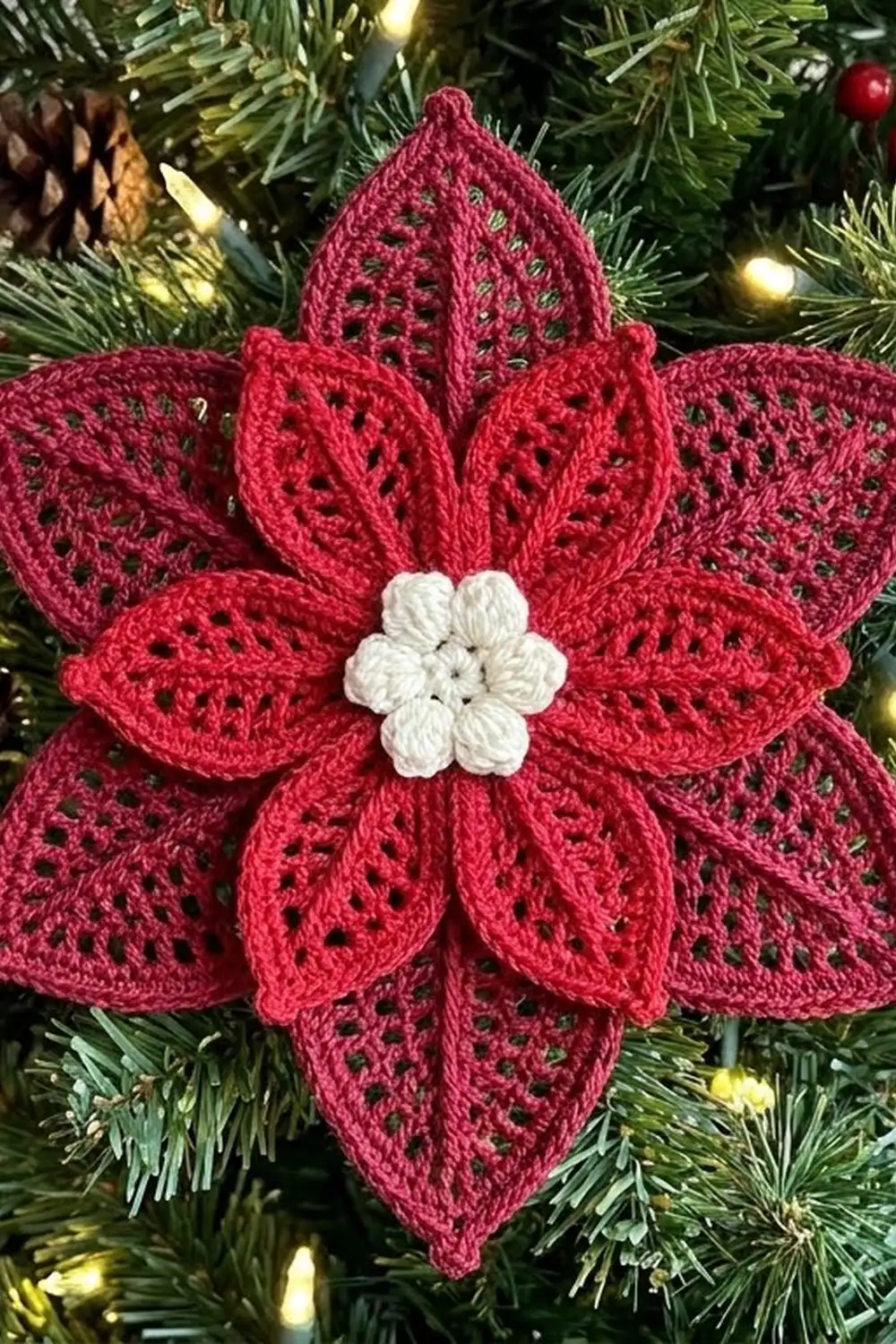 Deep red crocheted poinsettia with white center ornament displayed on an evergreen Christmas tree with warm lights.