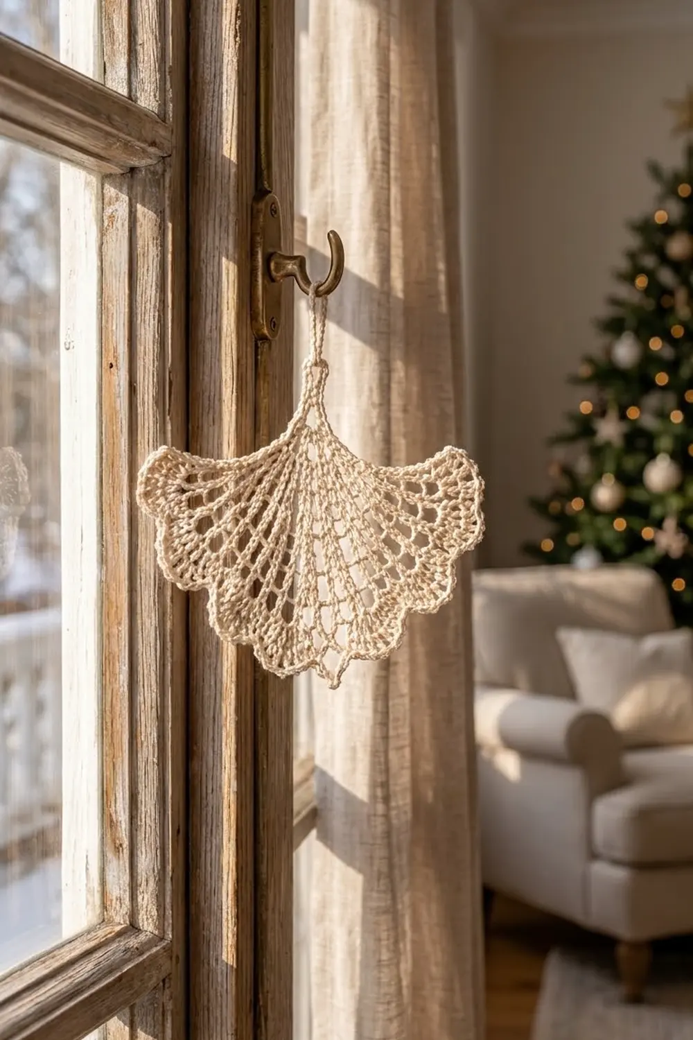 Delicate cream crocheted fan ornament hangs from brass hook on weathered wooden window frame with Christmas tree in background.