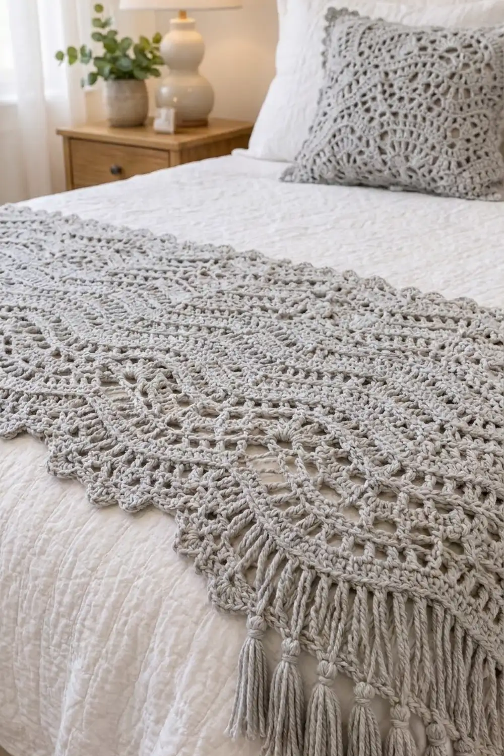Light gray crocheted blanket with delicate openwork lace and fringe styled on a bedroom bed.
