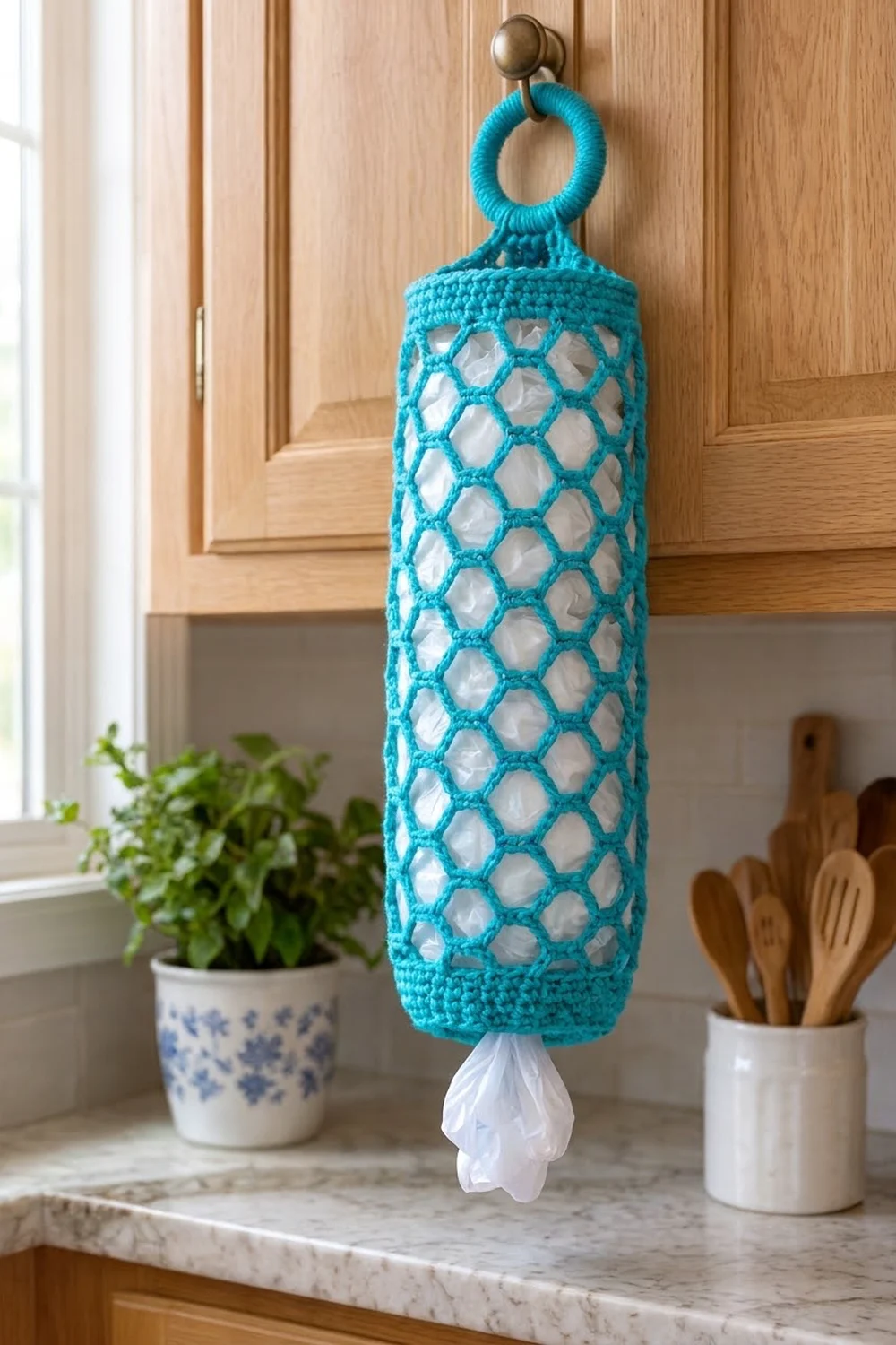 Turquoise crocheted mesh bag with diamond lattice pattern hanging from a brass knob on a light wooden kitchen cabinet.