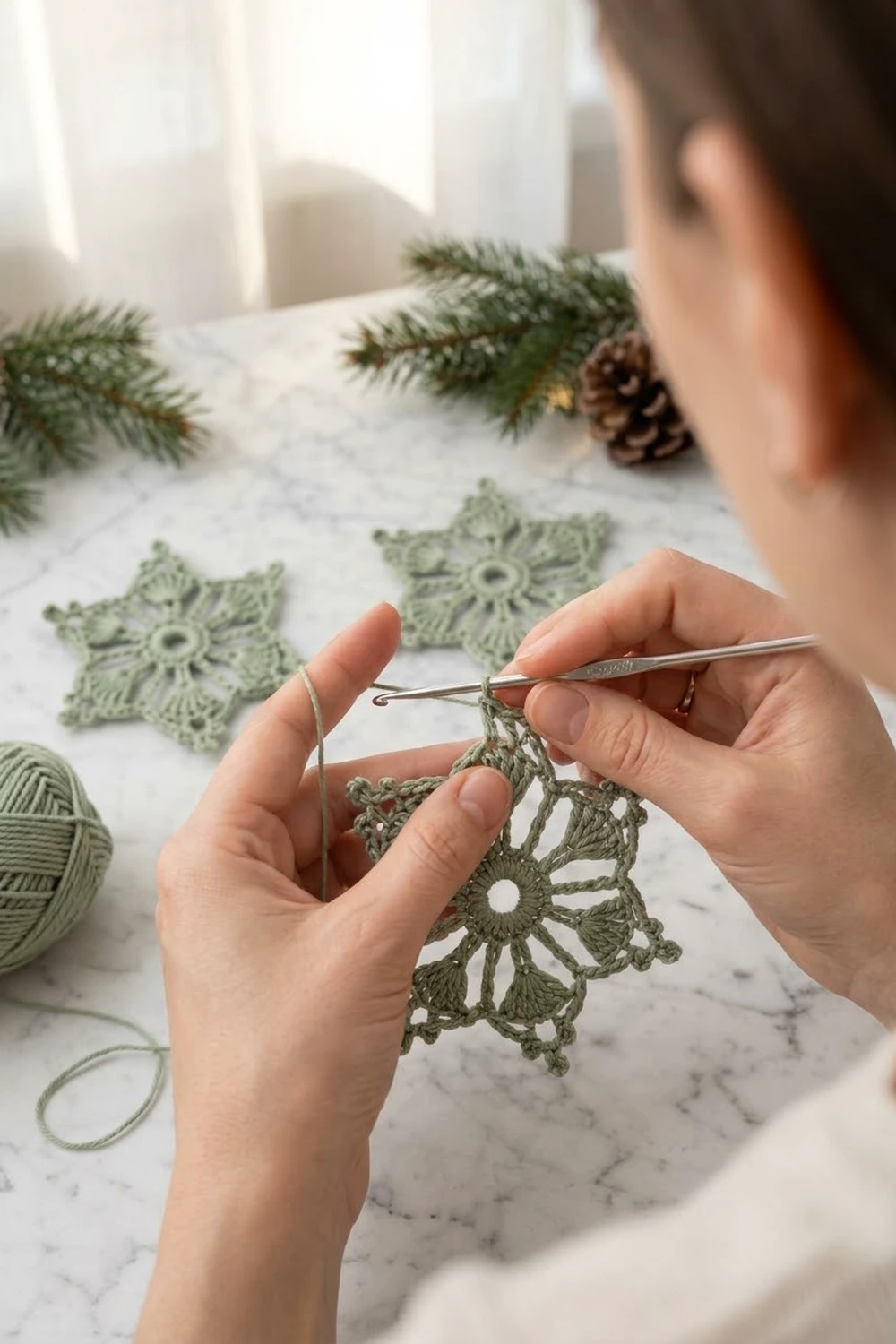 Person crocheting a delicate green snowflake ornament with a crochet hook on a marble table surrounded by evergreen branches and pinecones.