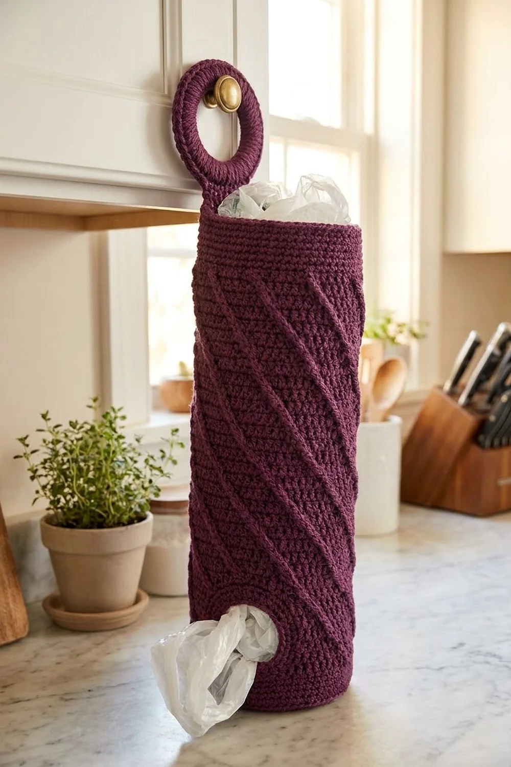 Deep burgundy crocheted bag with brass handle holds kitchen waste on bright countertop beside fresh potted herbs.