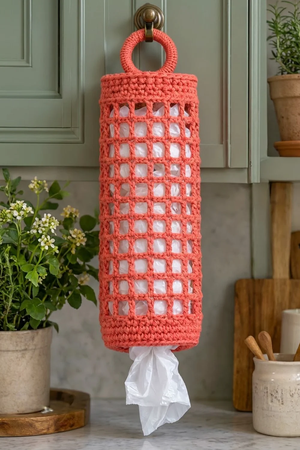 Coral crocheted plastic bag holder with openwork grid pattern hanging from a brass ring on a kitchen door.