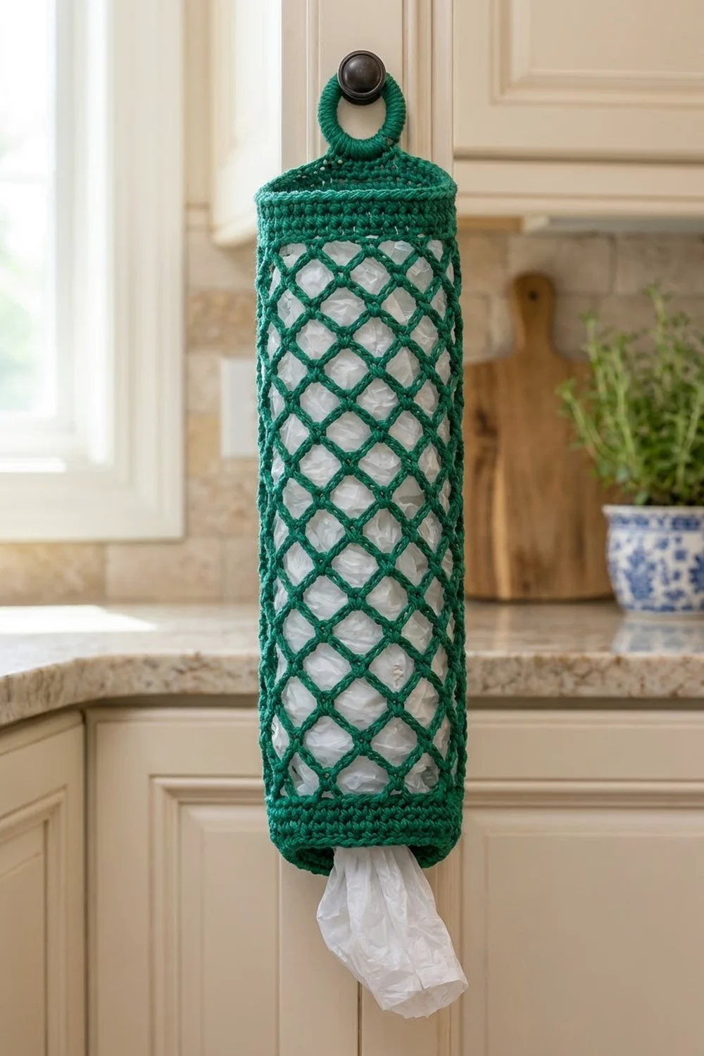 Teal crocheted plastic bag dispenser with diamond lattice pattern hanging from kitchen cabinet knob.