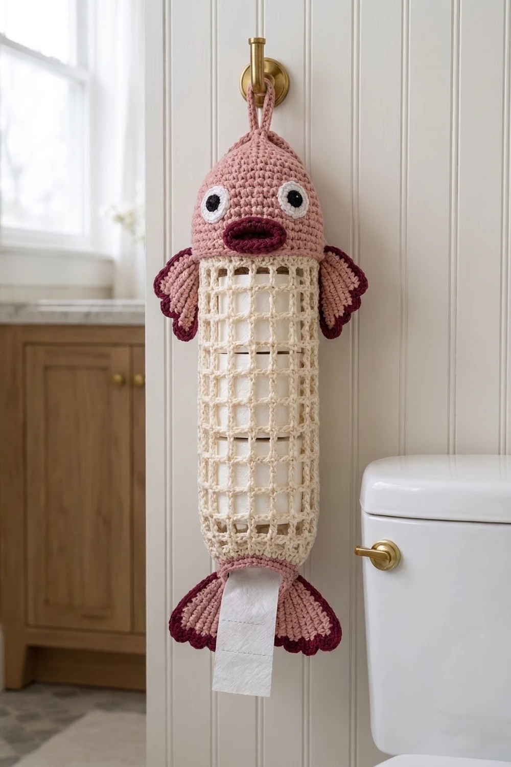 Whimsical crocheted fish toilet paper holder with pink head, cream body, and burgundy fins on bathroom wall.