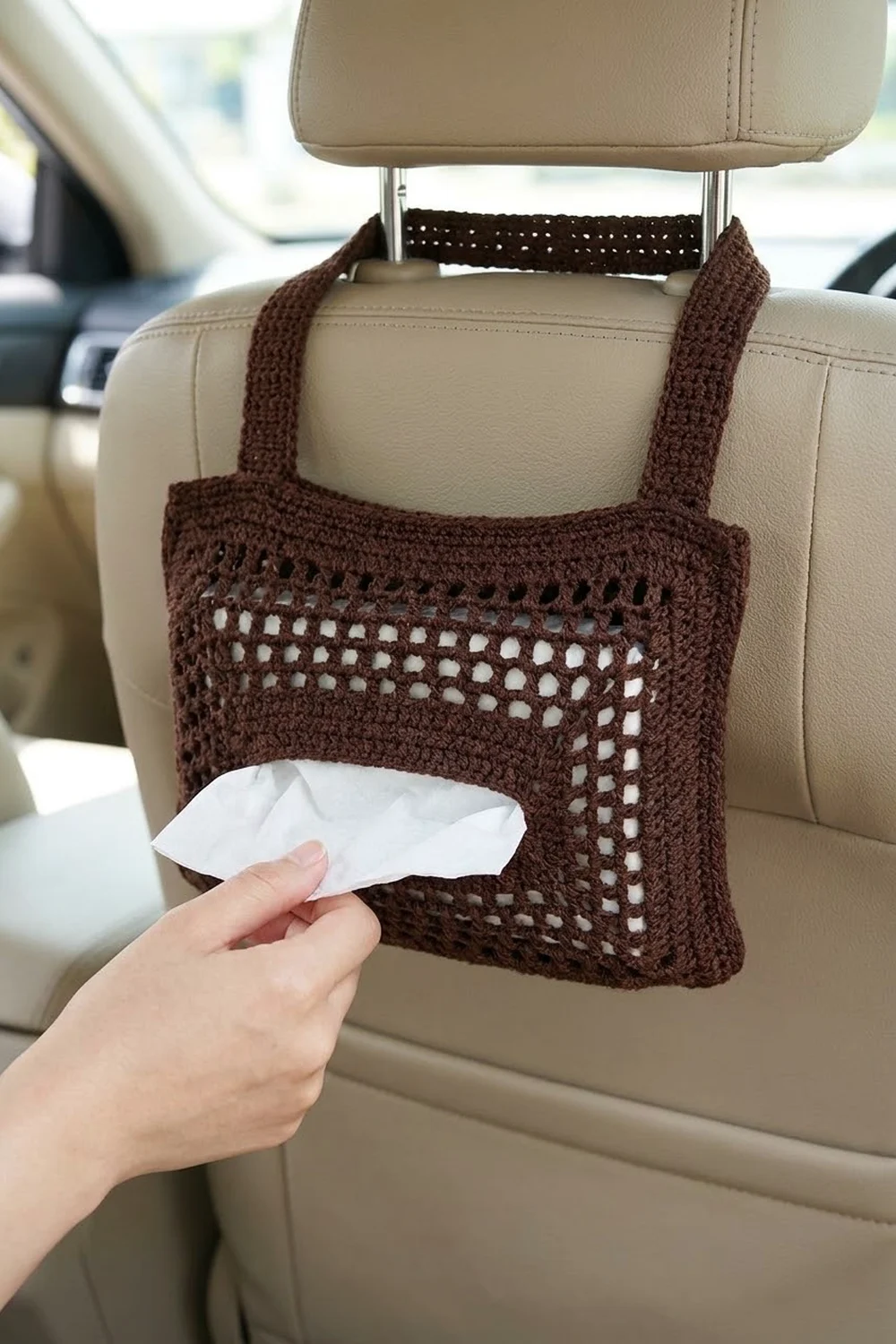 Brown crocheted car organizer with openwork pocket hanging from beige seat, dispensing white tissue inside a car.