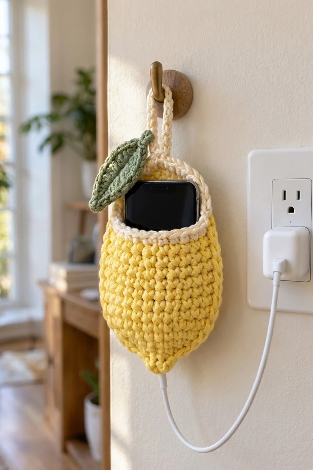 Yellow crocheted lemon-shaped phone holder with green leaf, mounted on cream wall beside outlet, holding black smartphone in minimalist home.