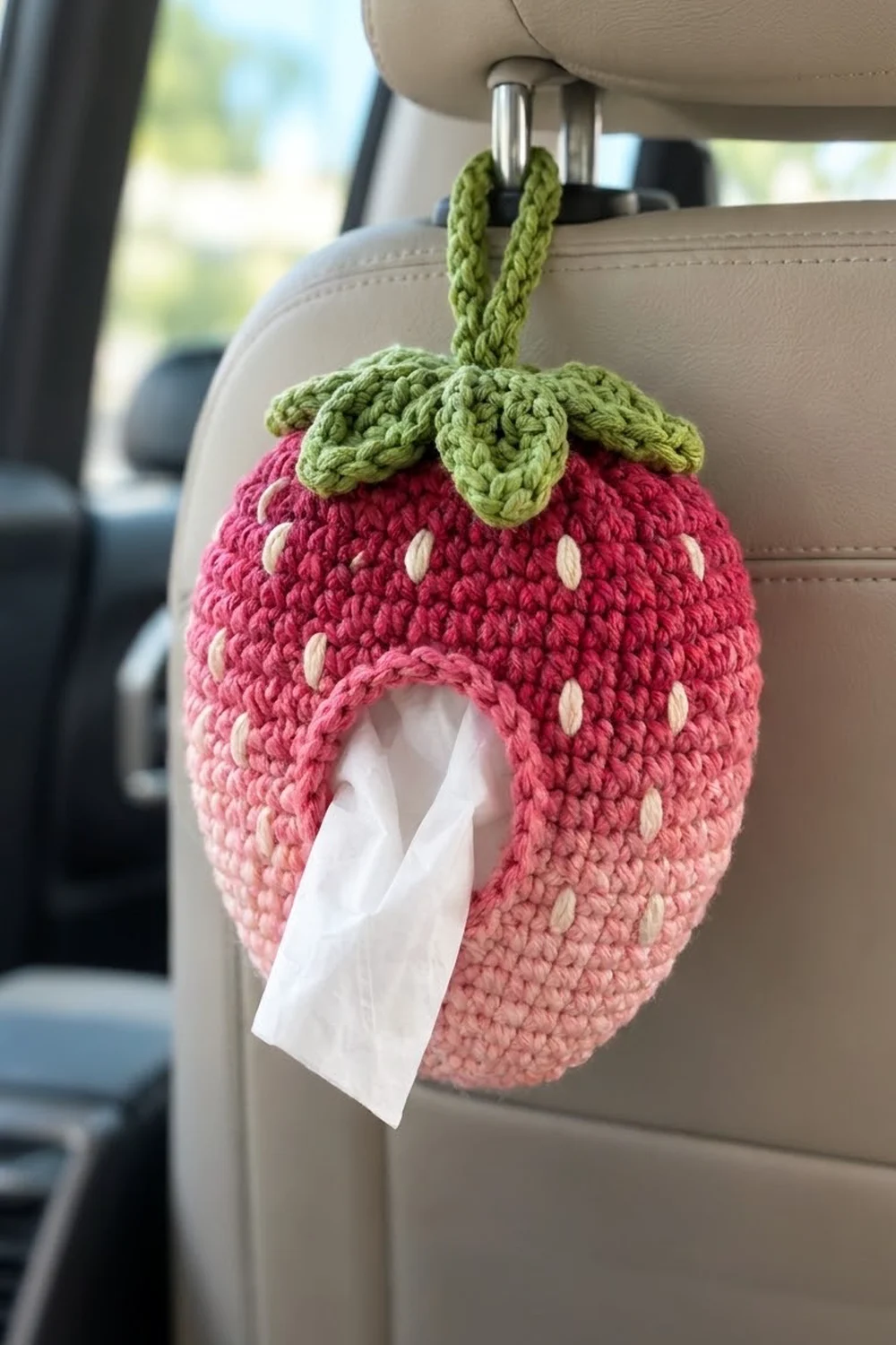 Crocheted pink strawberry with white seeds and green leaf crown hanging in a beige car interior.