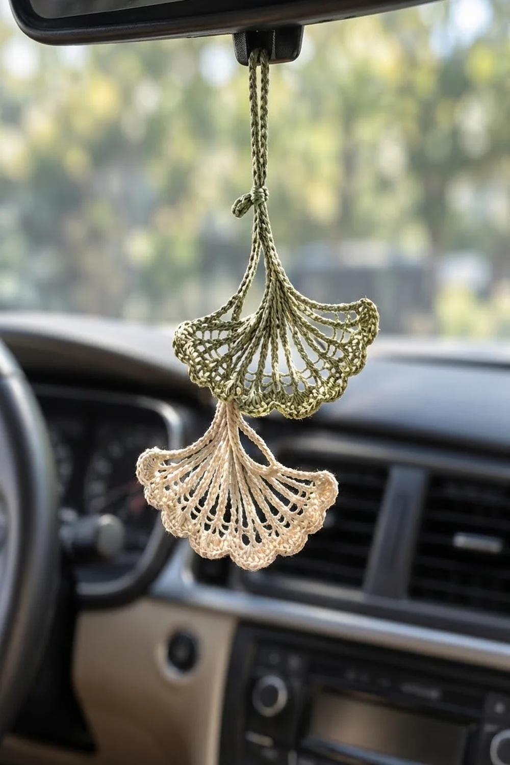 Two crocheted ginkgo leaves in sage green and cream dangle from a car's rearview mirror.