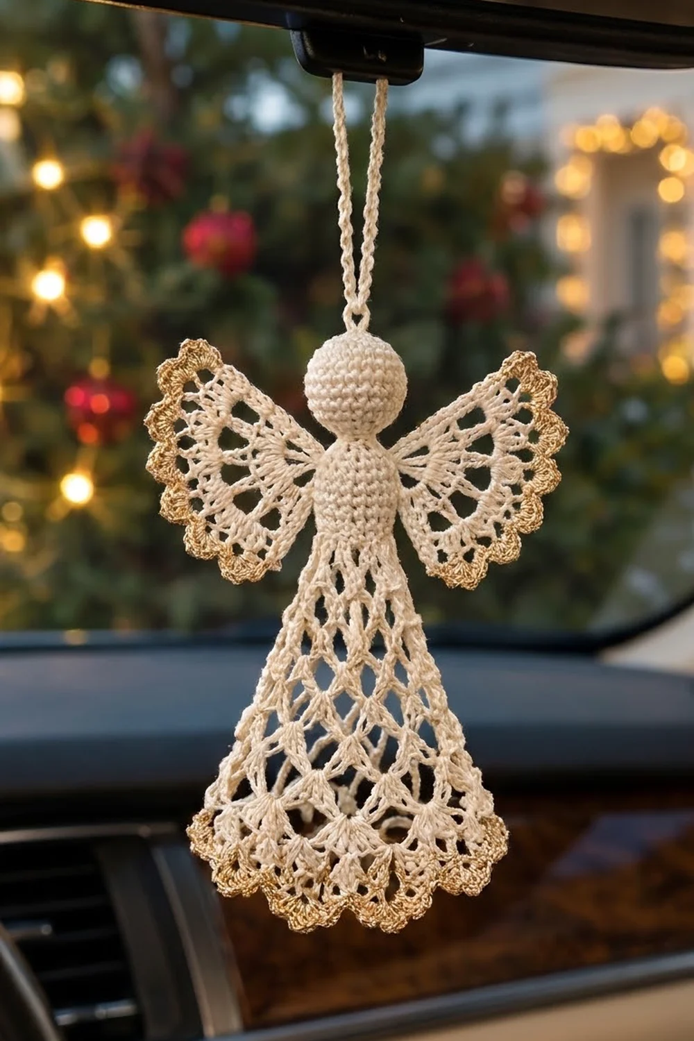 Cream and gold crocheted angel ornament with lace wings hanging from a car rearview mirror, Christmas wreath visible in background.