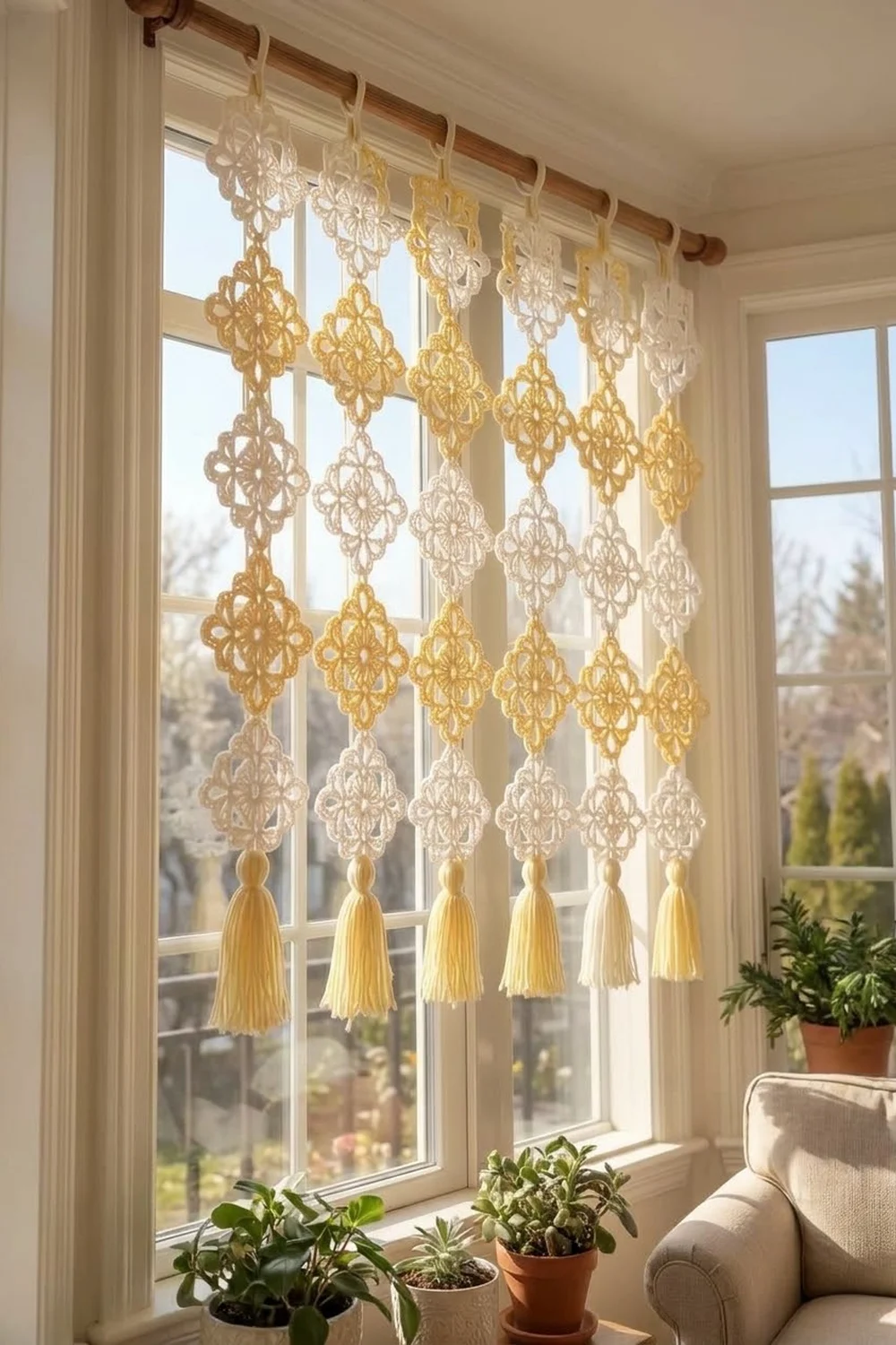 Golden and cream lace ornaments with tassels hang from a wooden rod across a sunlit window above potted plants.