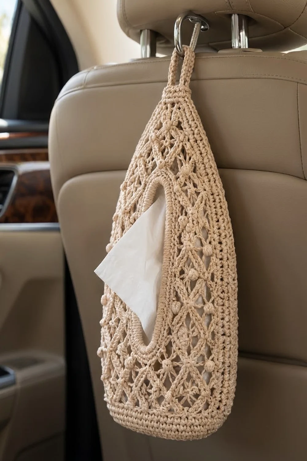 Beige crocheted car organizer with openwork lace pattern hanging from seat headrest, holding tissues.