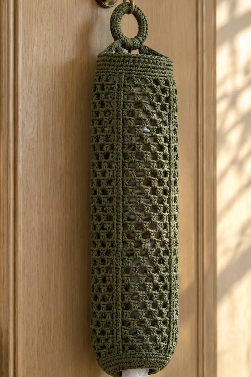 Sage-green crocheted hanging pouch with geometric openwork pattern against light wood paneling.