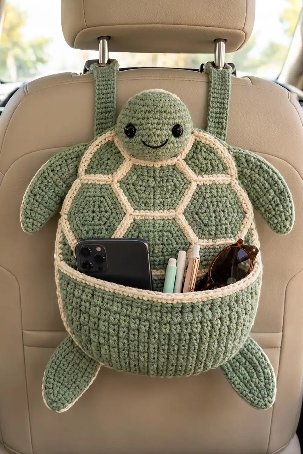 Sage green crocheted turtle organizer with cream shell hanging from beige car seat, holding smartphone and sunglasses in its pouch.