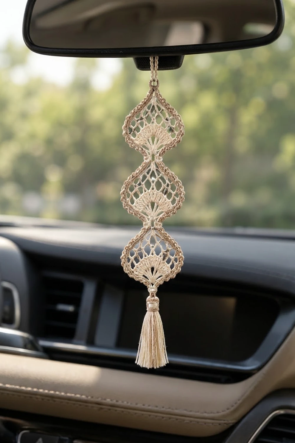 Beige crocheted ornament with three lace diamond shapes and tassel hanging from a rearview mirror in a car.
