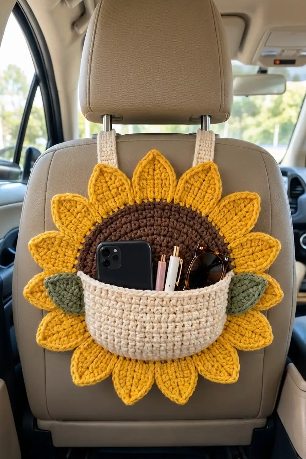 Crocheted sunflower organizer with yellow petals, brown center, and cream pocket holding phone and accessories on a beige car seat.