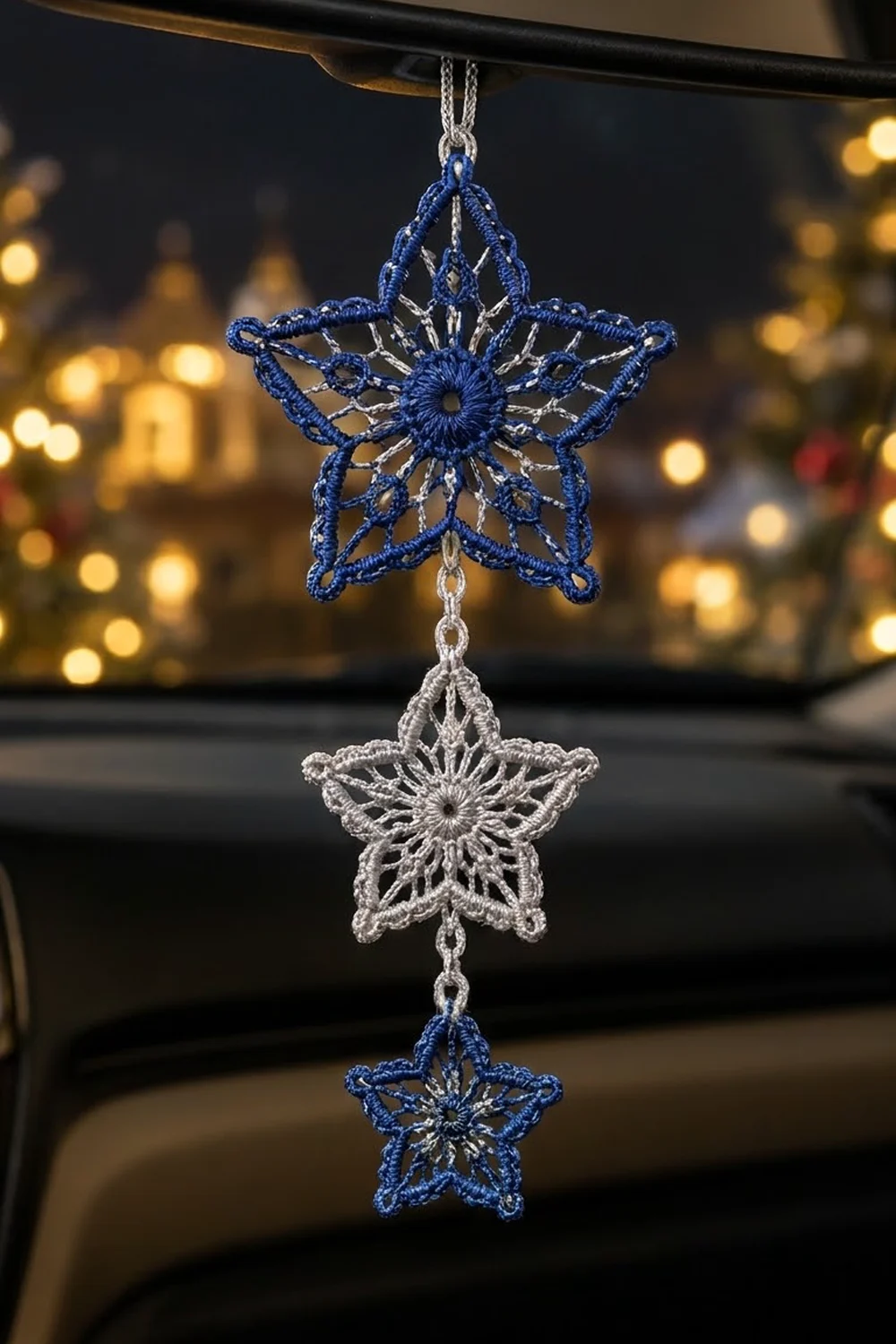 Three crocheted star ornaments in blue and silver hang from a chain on a car mirror, with festive golden lights blurred in the background.